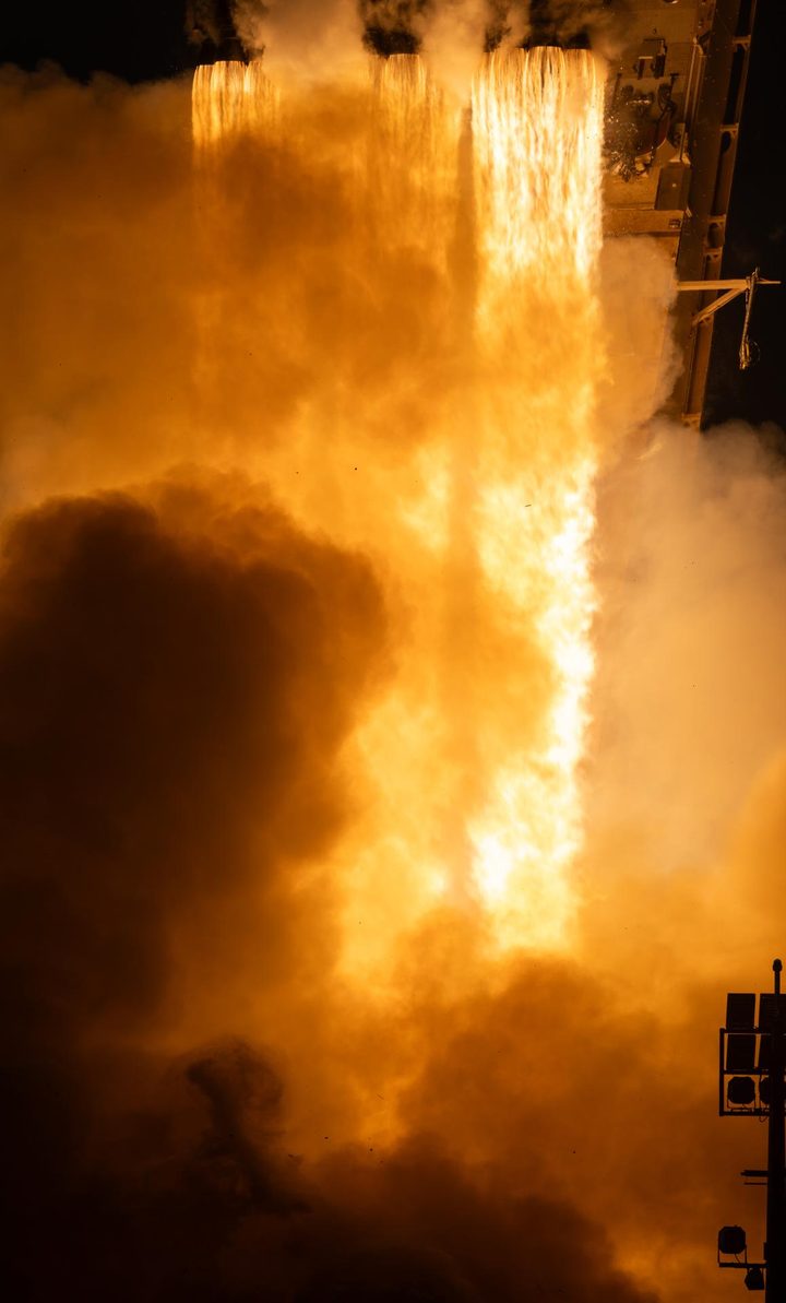 Bright orange flames and billowing smoke at liftoff, seen from beside the launch pad.