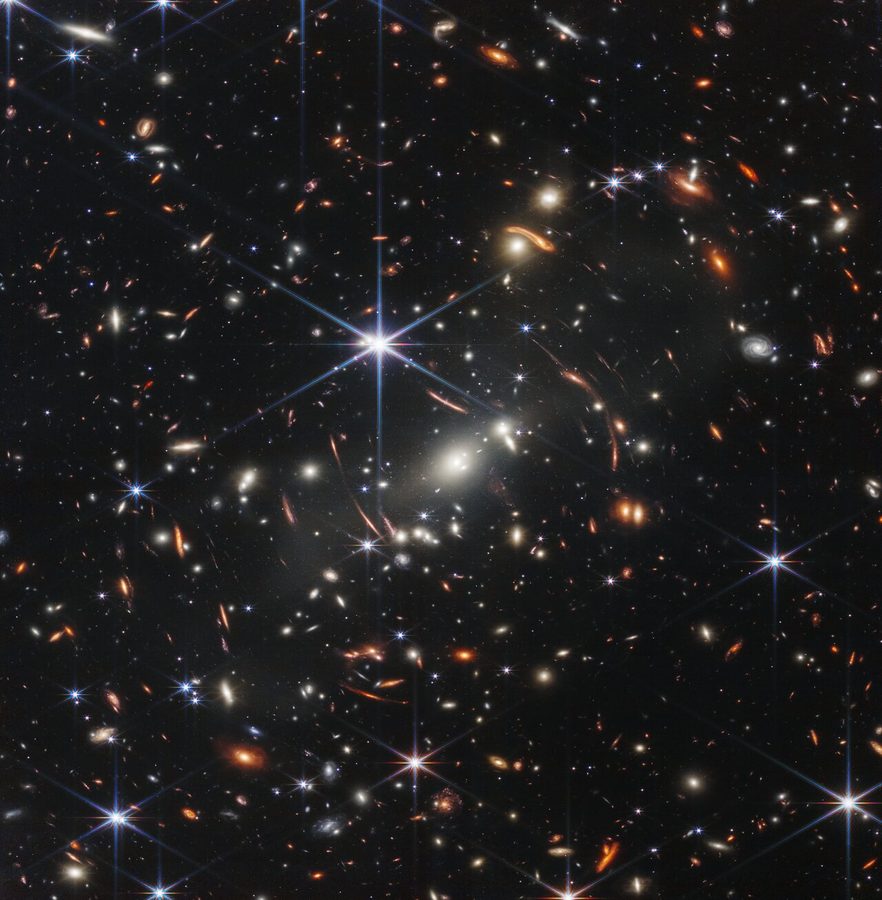 Thousands of galaxies of many shapes and colors; the bright galaxies of a foreground cluster are surrounded by thin, stretched arcs of more distant galaxies.